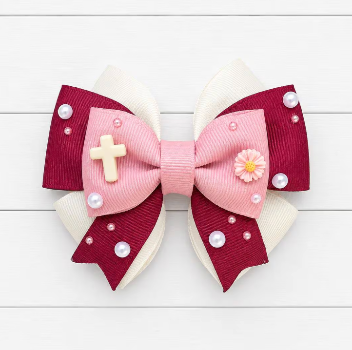 Hair bow with pink center, red outer layers, white accents, and small embellishments on a light wooden background. Cross charm on the left and flower charm on the right.