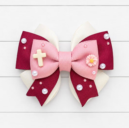 Hair bow with pink center, red outer layers, white accents, and small embellishments on a light wooden background. Cross charm on the left and flower charm on the right.