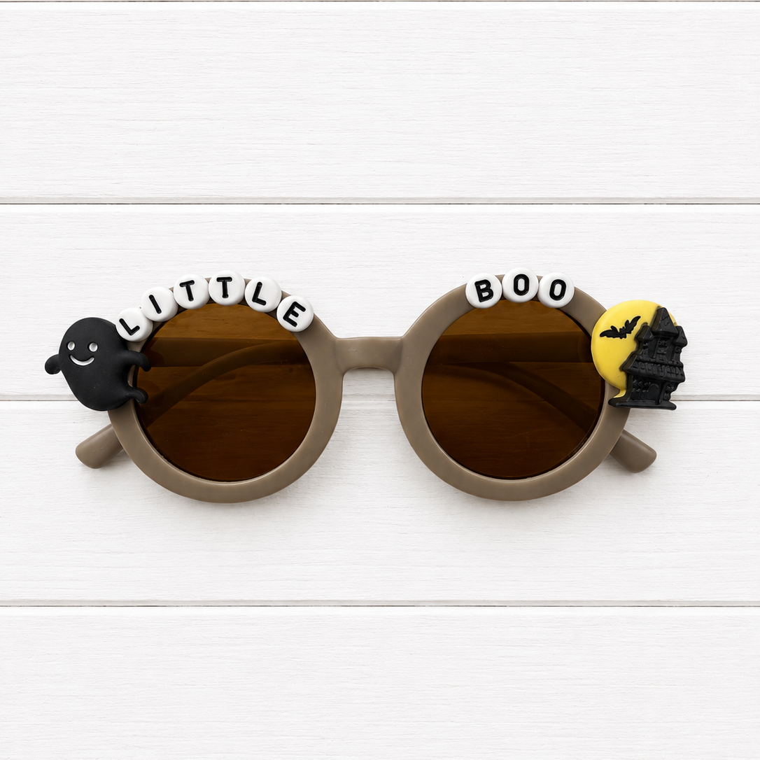 Round sunglasses with brown lenses and taupe frames, decorated with Halloween-themed charms including a black ghost, white letter beads spelling 'LITTLE BOO', and a yellow bat silhouette, shown on a white wood plank background.
