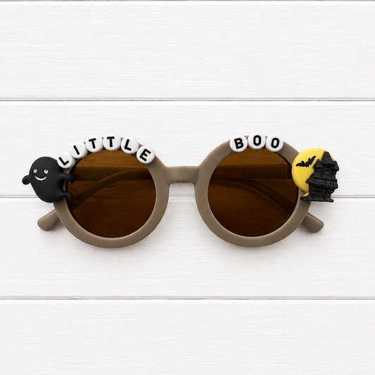 Round sunglasses with brown lenses and taupe frames, decorated with Halloween-themed charms including a black ghost, white letter beads spelling 'LITTLE BOO', and a yellow bat silhouette, shown on a white wood plank background.