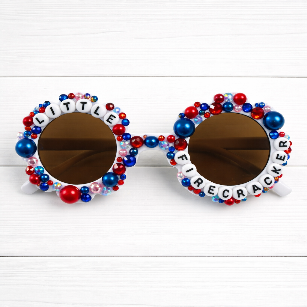 Kids flower white Sunglasses with red and blue rhinestones and pearls on frame and 'LITTLE FIRECRACKER' text on a white wooden background