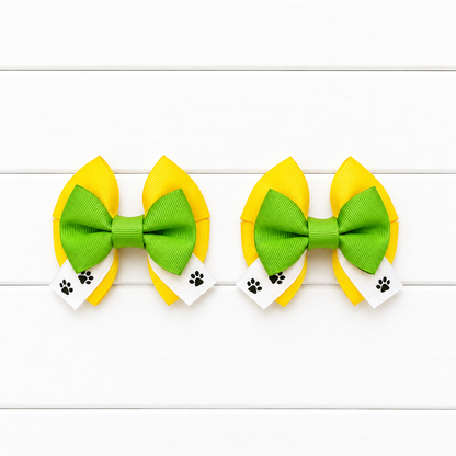 Two matching hair bows with yellow top and bottom, green middle bow, and white tails with paw prints on the ribbon