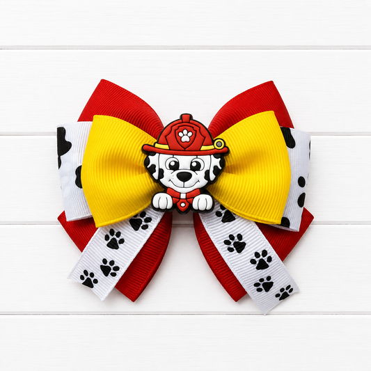 A children's hair bow featuring a red and yellow grosgrain ribbon loop, white ribbon loops with black paw prints, and a central Paw Patrol firefighter dalmatian character charm.