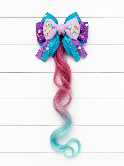 A mermaid-themed hair bow accessory with a purple and blue glitter bow, small decorative charms, and a long wavy synthetic hair extension in pink fading to light blue, displayed on a white wood plank background.