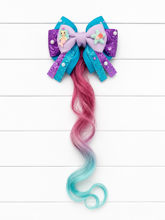 A mermaid-themed hair bow accessory with a purple and blue glitter bow, small decorative charms, and a long wavy synthetic hair extension in pink fading to light blue, displayed on a white wood plank background.