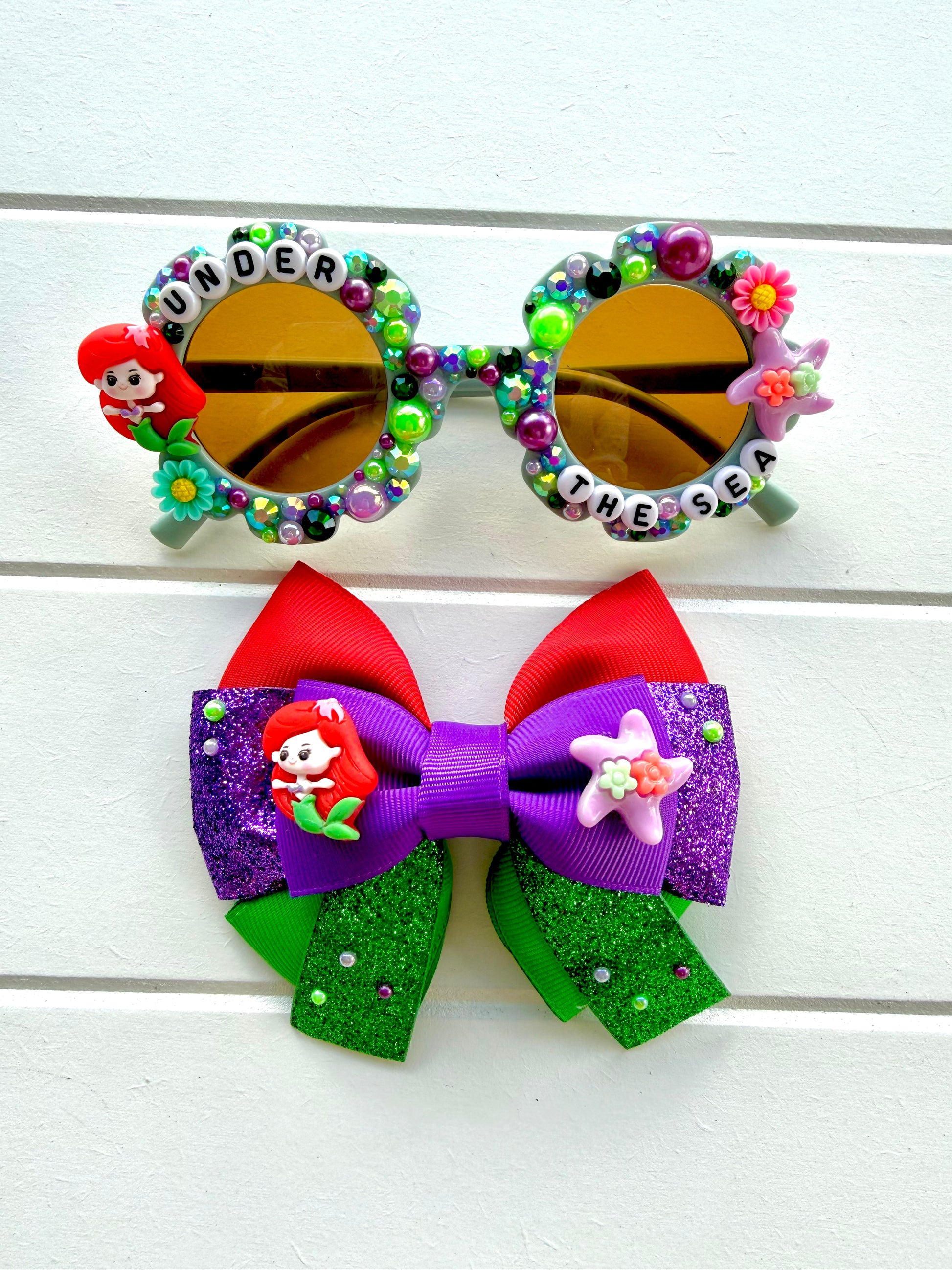 Matching sunglasses and bow. Kids green flower sunglasses with Ariel charm on left and starfish charm on right. "UNDER THE SEA" text, green and purple pearls and rhinestones and flowers outline the frames. The bow features red, green and purple ribbon with Ariel and starfish charms in the middle.