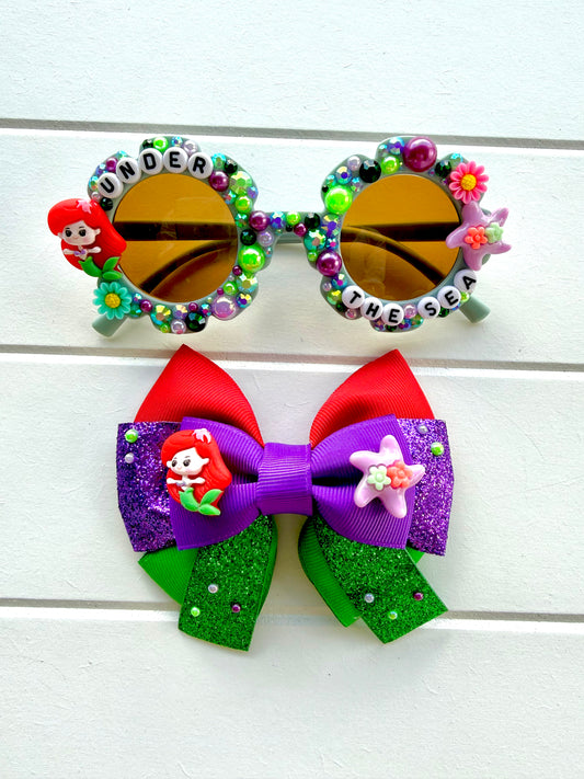 Matching sunglasses and bow. Kids green flower sunglasses with Ariel charm on left and starfish charm on right. "UNDER THE SEA" text, green and purple pearls and rhinestones and flowers outline the frames. The bow features red, green and purple ribbon with Ariel and starfish charms in the middle.