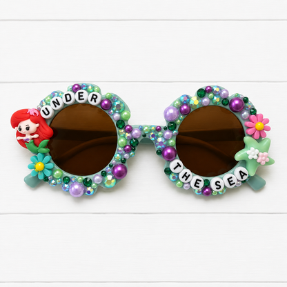 Kids green blue flower sunglasses with mermaid and starfish charms on either side and flowers, rhinestones, and pearls decorating the frames. "UNDER THE SEA" text outlines the frames.
