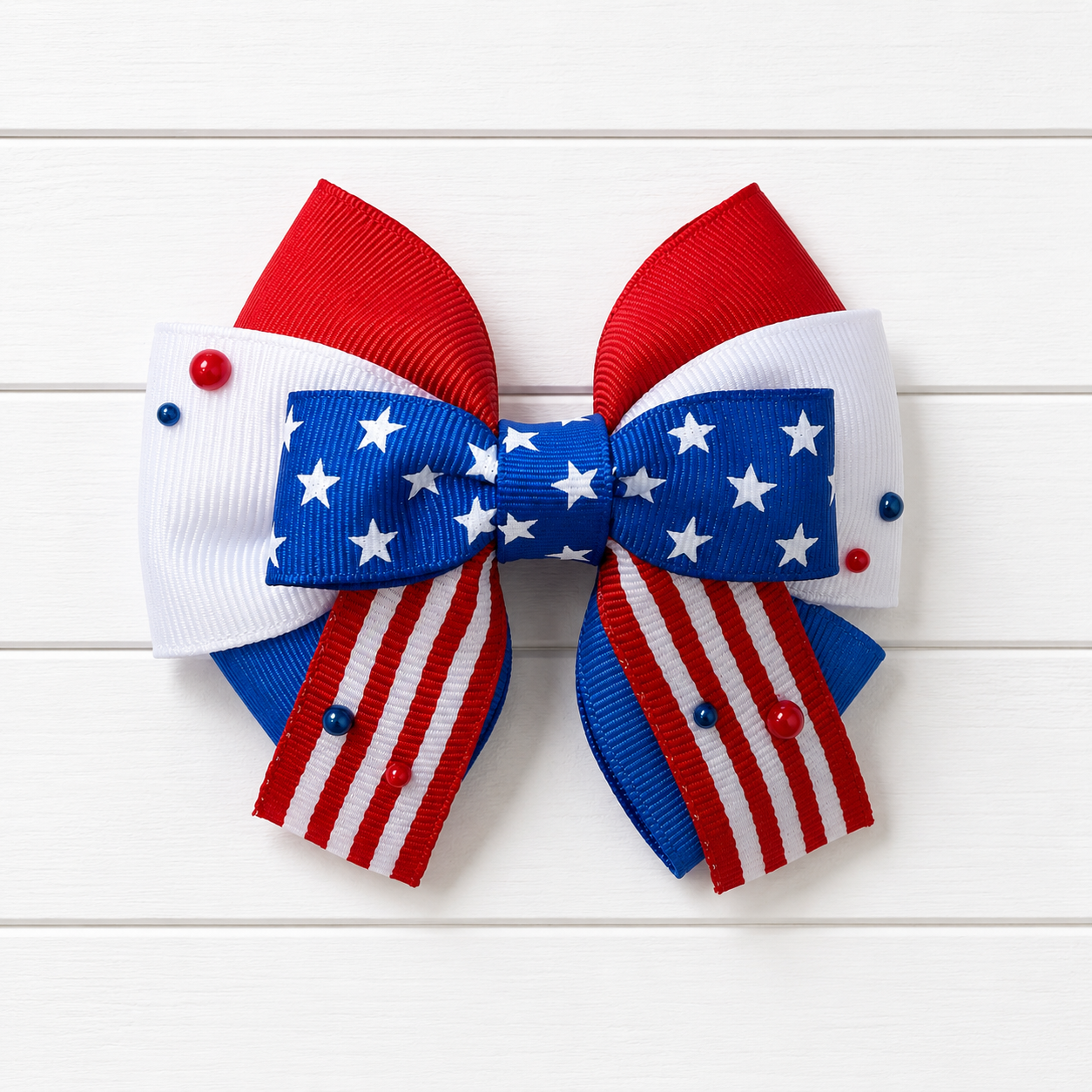 Girls hair bow with red white and blue colors and stars in the middle. Featuring red and white striped ribbon at the bottom with a couple of red and blue pearl embellishments scattered throughout.