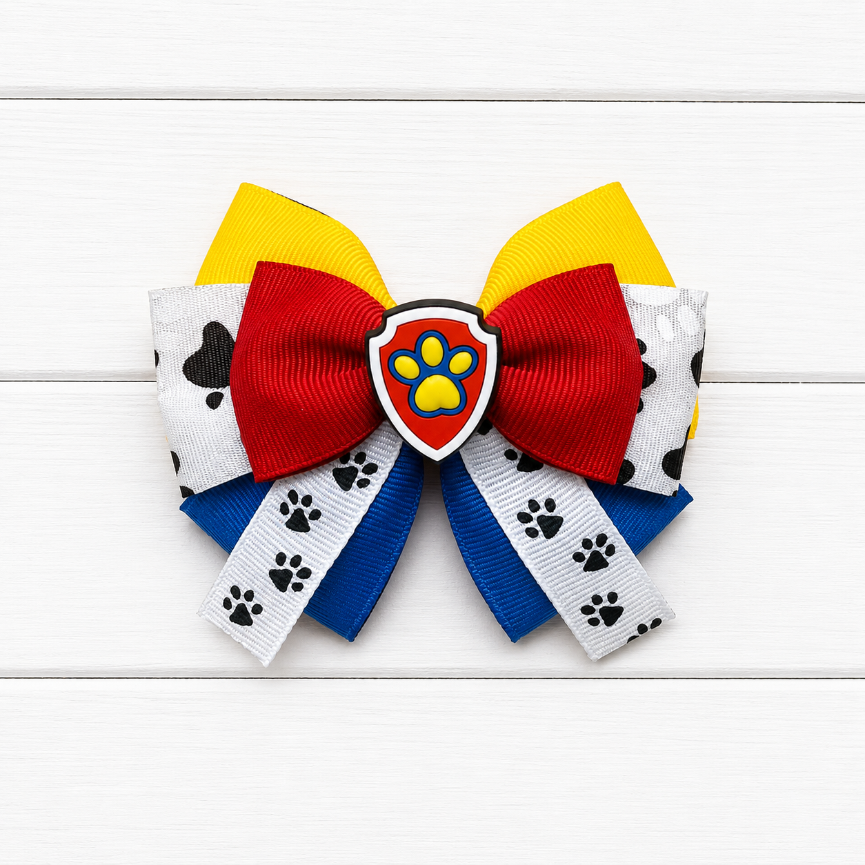 A girl's hair bow featuring red, yellow, blue, and white ribbons with black paw prints, centered with a Paw Patrol shield emblem.