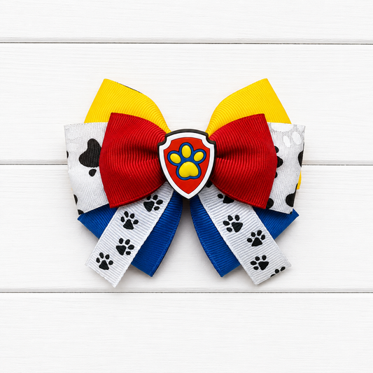 A girl's hair bow featuring red, yellow, blue, and white ribbons with black paw prints, centered with a Paw Patrol shield emblem.