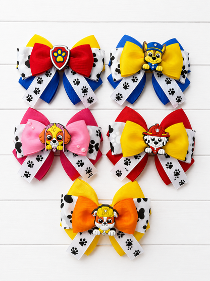 Six colorful Paw Patrol hair bows arranged on a white wood background. Each bow features a large color-blocked top section with red, yellow, blue, pink, or orange, a Paw Patrol character charm in the center, and white ribbon tails printed with black paw prints.
