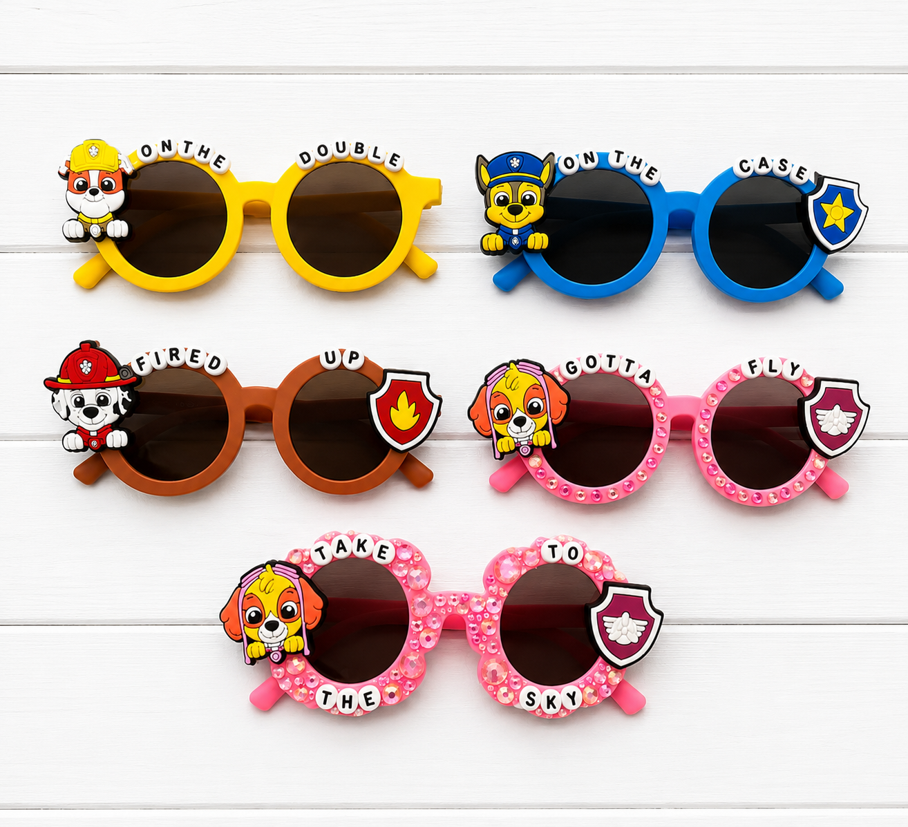 Six pairs of children's sunglasses arranged in two rows on a white wood background. The sunglasses feature colorful frames in yellow, blue, brown, and pink, each decorated with a Paw Patrol character charm and text labels such as 'ON THE DOUBLE', 'ON THE CASE', 'FIRED UP', 'GOTTA FLY', 'TAKE TO THE SKY'.