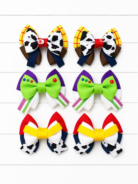Six colorful fabric hair bows arranged in two columns on a white wood plank background. The bows feature playful character-inspired designs, including cow print, plaid, green with purple and red accents, and red, yellow, white, and black-and-white cow print patterns.