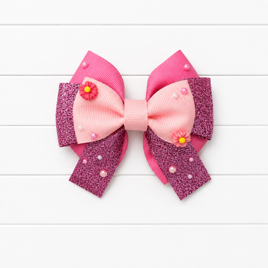 A pink hair bow featuring a center light pink grosgrain ribbon loop, a darker pink glitter loop, and two magenta glitter loops with small pink flowers and pearl embellishments, displayed on a white wood plank background.