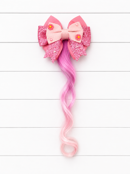 A pink hair bow accessory with glittery pink and light pink fabric, two small yellow flower embellishments, and a long wavy pink synthetic hair tail, displayed against a white wood plank background.