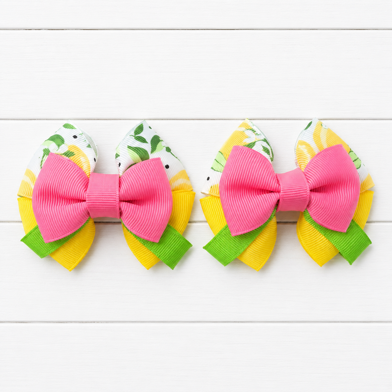 Two matching hair bows arranged side by side on a white wood plank background. Each bow has a large pink grosgrain ribbon center, yellow and green patterned fabric loops.