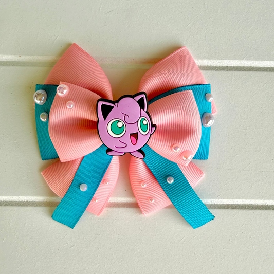 A pink and teal grosgrain ribbon hair bow featuring a Jigglypuff character charm in the center, decorated with small pearl-like beads, shown against a light beige paneled wall.