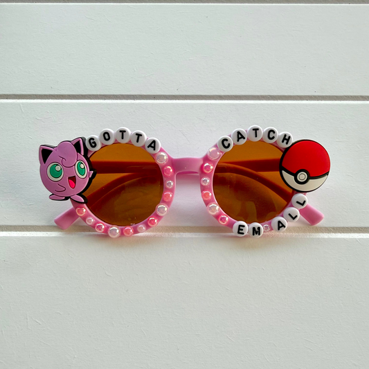 Pink novelty sunglasses with Jigglypuff and Pokéball graphics, letter beads spelling 'GOTTA CATCH EM ALL', and rhinestone trim, shown on a white paneled background.