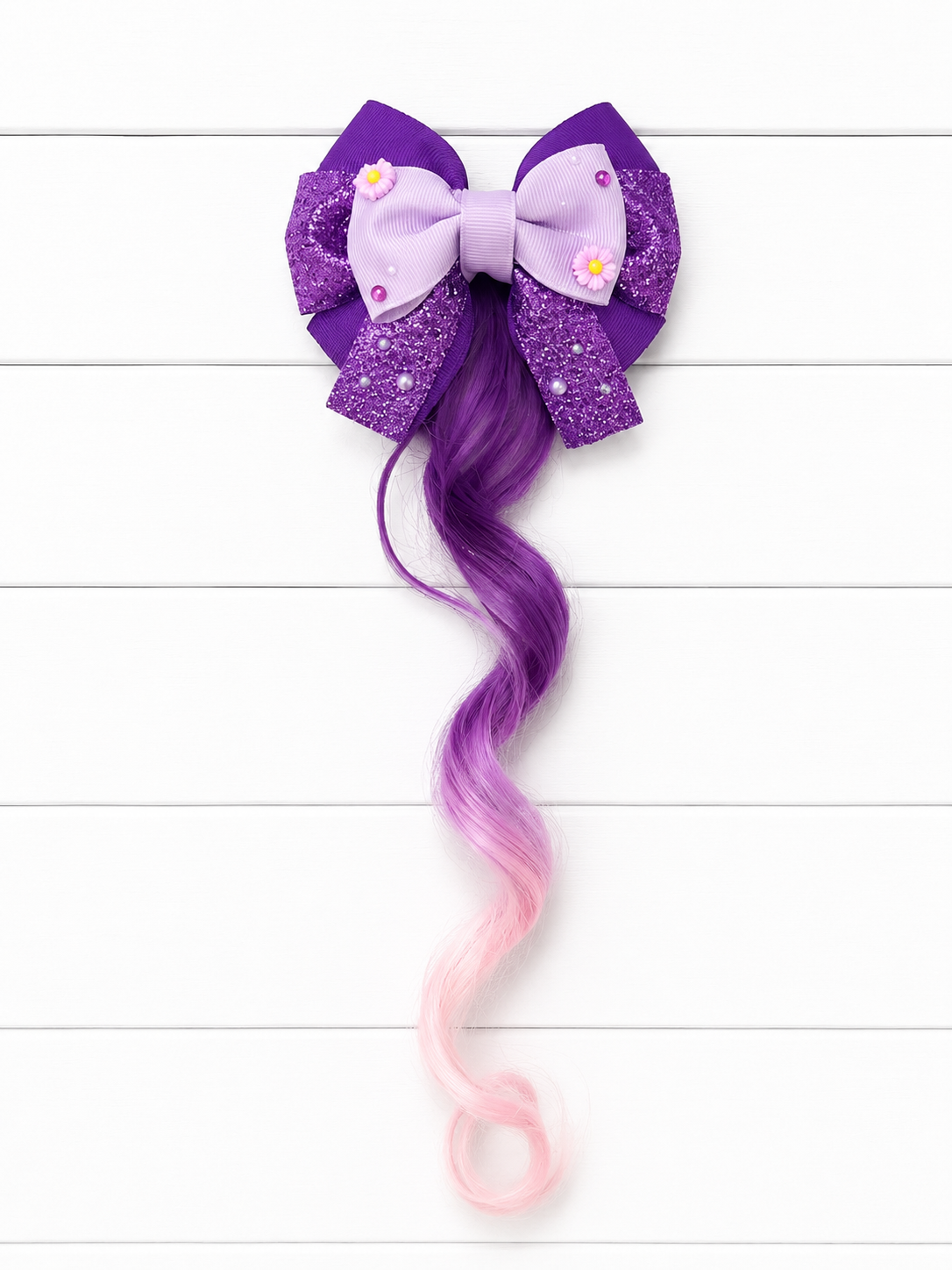 A purple and pink ombre wavy hair extension styled in a large bow with glittery purple side wings and small daisy embellishments, displayed against a white wood plank background.