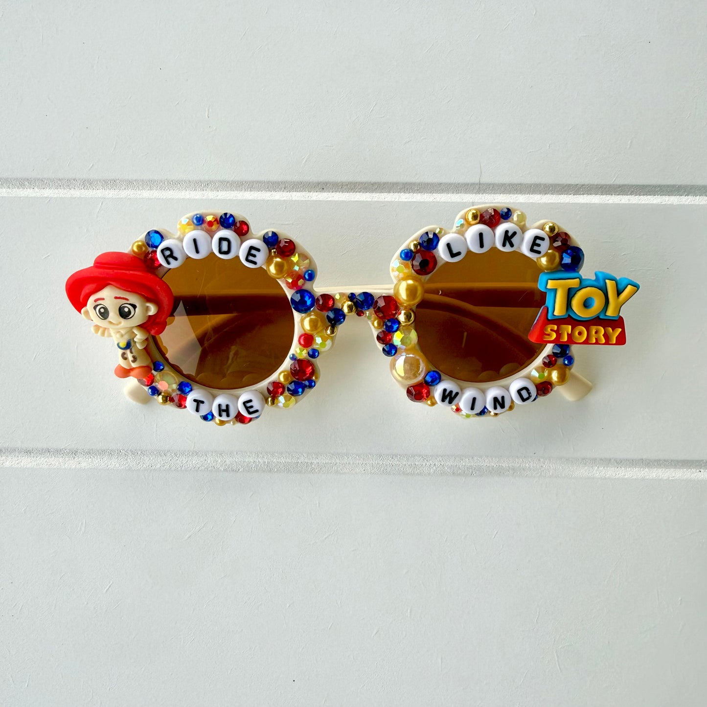 Kids cream flower sunglasses with Jessie charm on left and Toy Story charm on right. Red, yellow, and blue pearls and rhinestones and "RIDE LIKE THE WIND" text outlines the frames.
