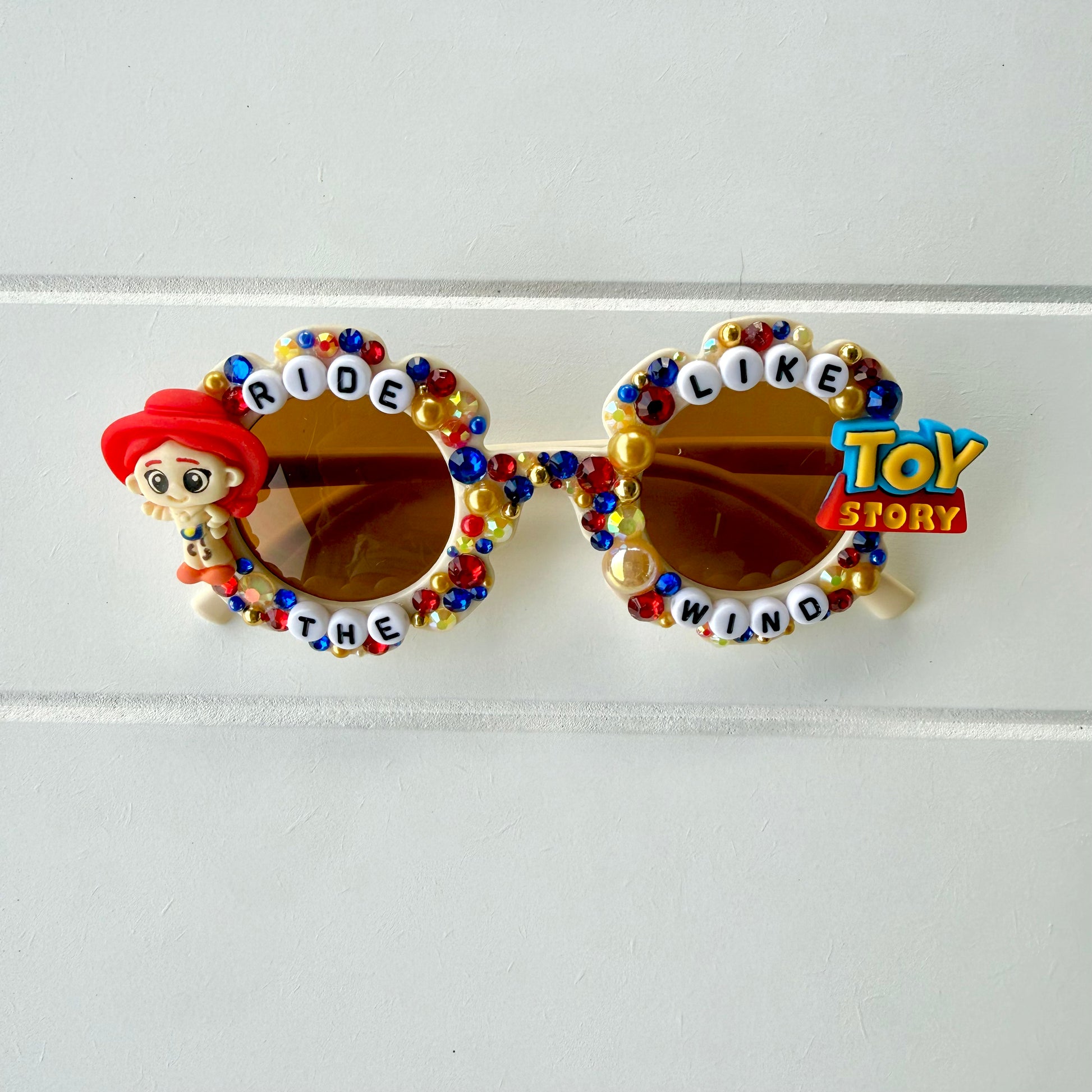 Kids cream flower sunglasses with Jessie charm on left and Toy Story charm on right. Red, yellow, and blue pearls and rhinestones and "RIDE LIKE THE WIND" text outlines the frames.