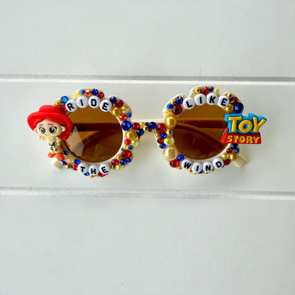 Kids cream flower sunglasses with Jessie charm on left and Toy Story charm on right. Red, yellow, and blue pearls and rhinestones and "RIDE LIKE THE WIND" text outlines the frames.