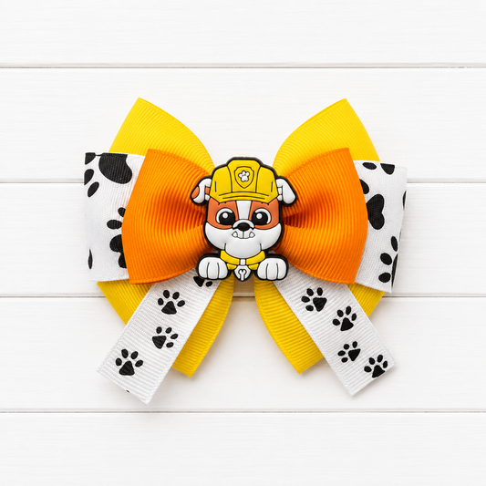 A pet hair bow featuring a large orange center loop, two yellow side loops, white ribbon tails with black paw prints, and a Rubble character charm on top, displayed on a white wood background.