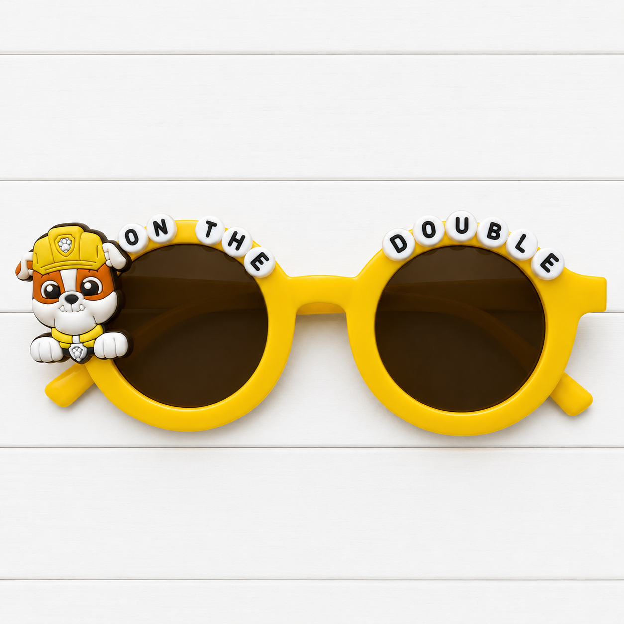Kids' yellow sunglasses with Rubble Paw Patrol charm and "ON THE DOUBLE" text outlining the frames