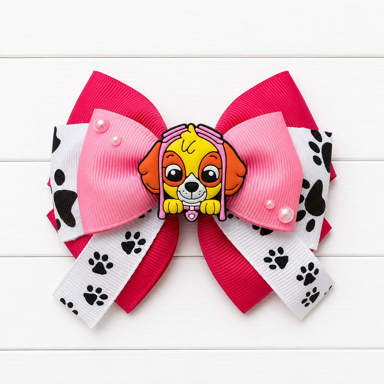 A Paw Patrol themed hair bow featuring Skye in the center, layered with hot pink, light pink, white, and black paw print ribbons, accented with two small pearl-like beads.