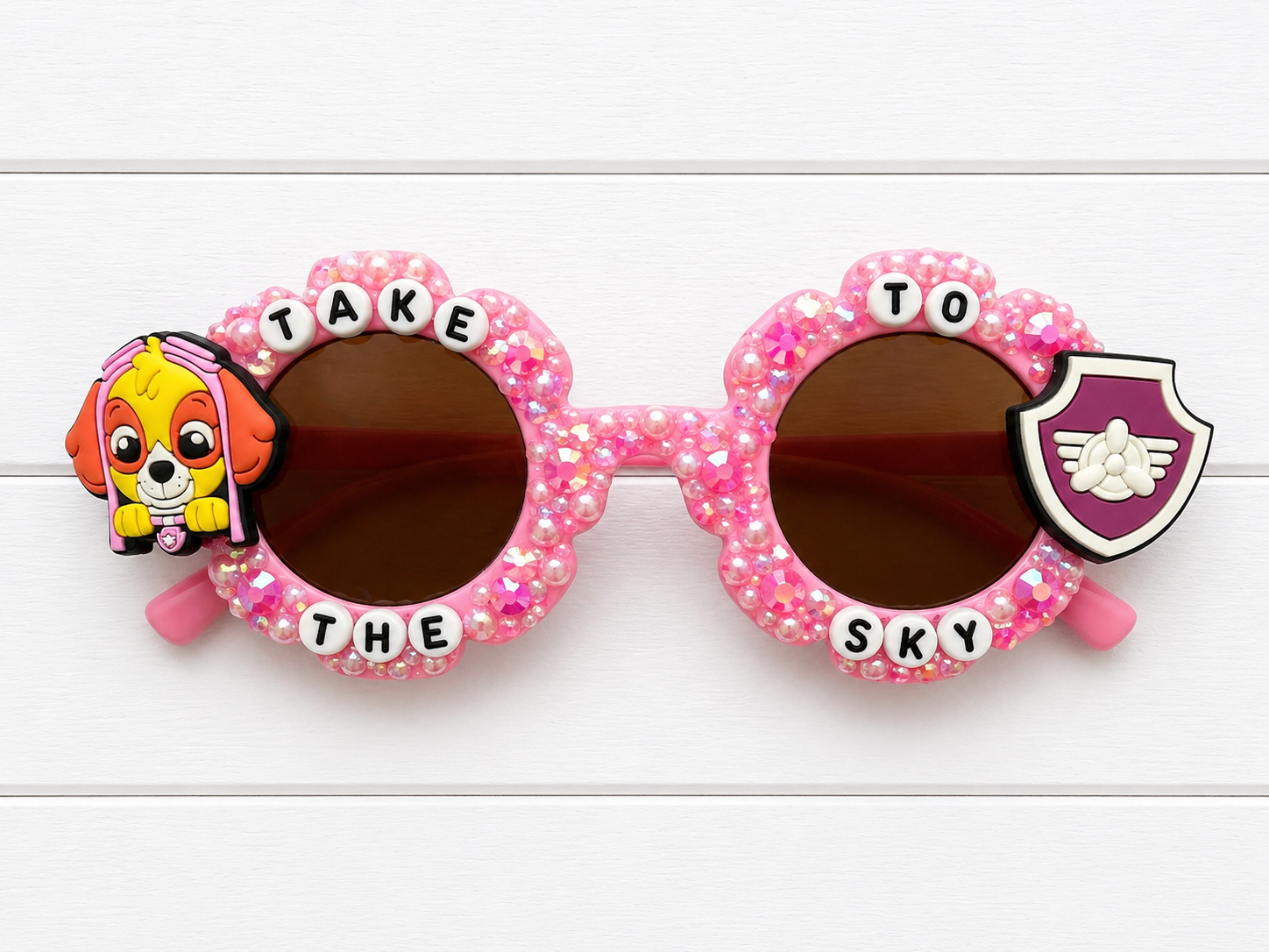 Pink flower kid's sunglasses with pink rhinestone and pearl embellishments and Skye Paw Patrol charms on either side. "TAKE TO THE SKY" text outlines the frames.