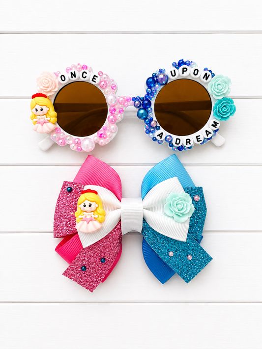 Matching sunglasses and bow themed around Aurora. Kids Sunglasses are white flower frames with Aurora charm on left and blue flowers on the right. "ONCE UPON A DREAM" letters and pink and blue pearls and rhinestones outline the frames. The Bow features pink and blue ribbon and an Aurora and rose charm in the middle.
