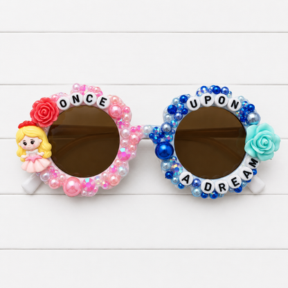 Kids white flower sunglasses with a pink princess and pink and blue rose charms on either side. Pink pearls and rhinestones on the left side and blue pearls and rhinestones on the right side decorating the frames. "ONCE UPON A DREAM" text outlines the frames.