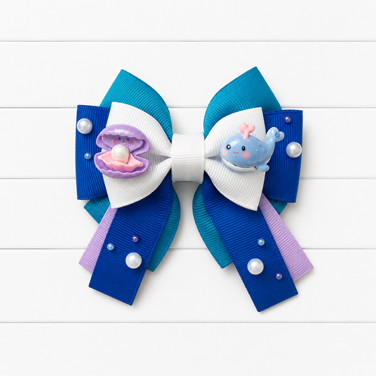 A blue, purple, and white hair bow featuring a clam and whale charms, embellished with small pearl-like beads, set against a white wood plank background.