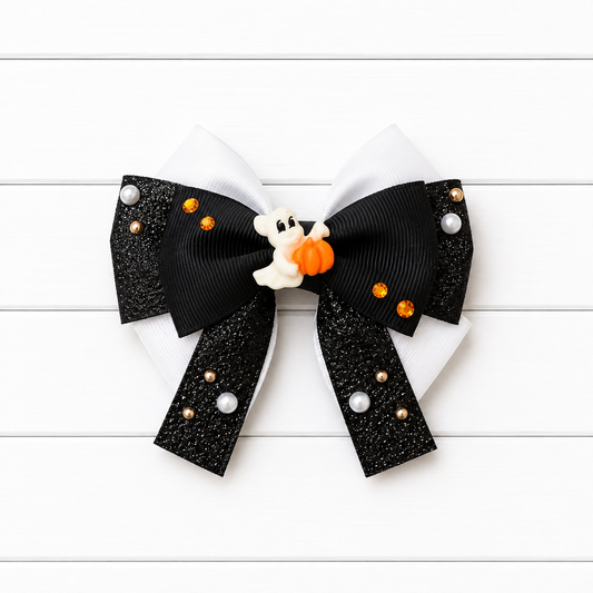 A Halloween-themed hair bow featuring black glitter ribbon tails, white grosgrain loops, small pearl-like and orange metallic embellishments, and a small white ghost holding an orange pumpkin at the center, displayed on a white wood plank background.