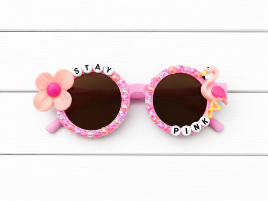 Kids round pink sunglasses with pink flower on left and flamingo on right. "STAY PINK" text outlines the frames and pink rhinestones and pearls outlines the frames.