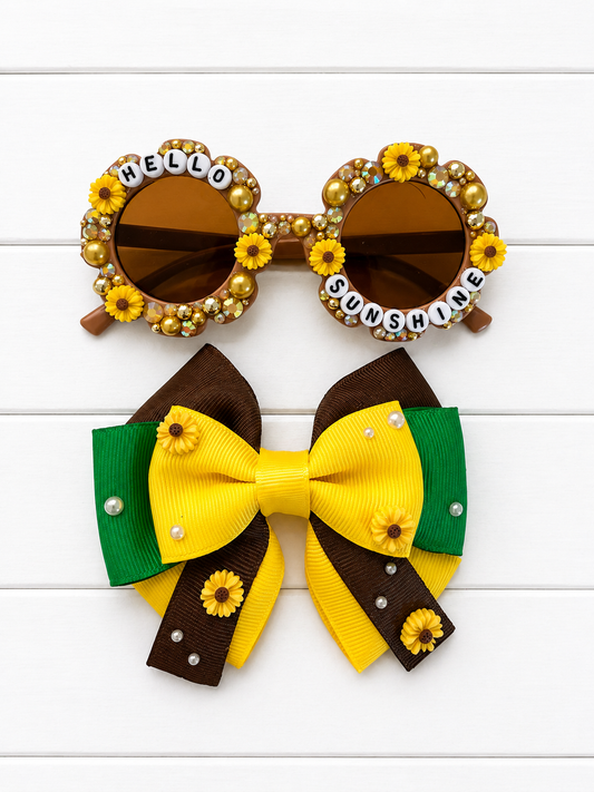 A styled flat-lay of a matching hair accessory set featuring a large bow and a pair of sunglasses. The bow is made of yellow, brown, and green grosgrain ribbon with small sunflower embellishments and pearl accents. The sunglasses match with brown flower frames covered with sunflower charms and gold and yellow rhinestones and pearls. "HELLO SUNSHINE" text outlines the frames.
