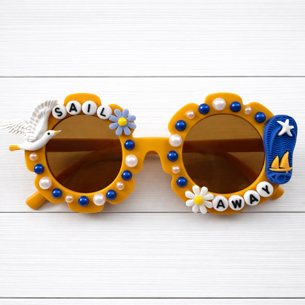 Kids dark yellow flower sunglasses with a seagull charm on left and flip flop charm on the right and "Sail Away" text outlining the frames. There are blue and white pearls outlining the frames.