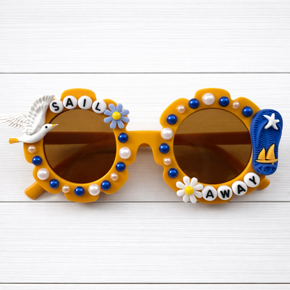Kids dark yellow flower sunglasses with a seagull charm on left and flip flop charm on the right and "Sail Away" text outlining the frames. There are blue and white pearls outlining the frames.