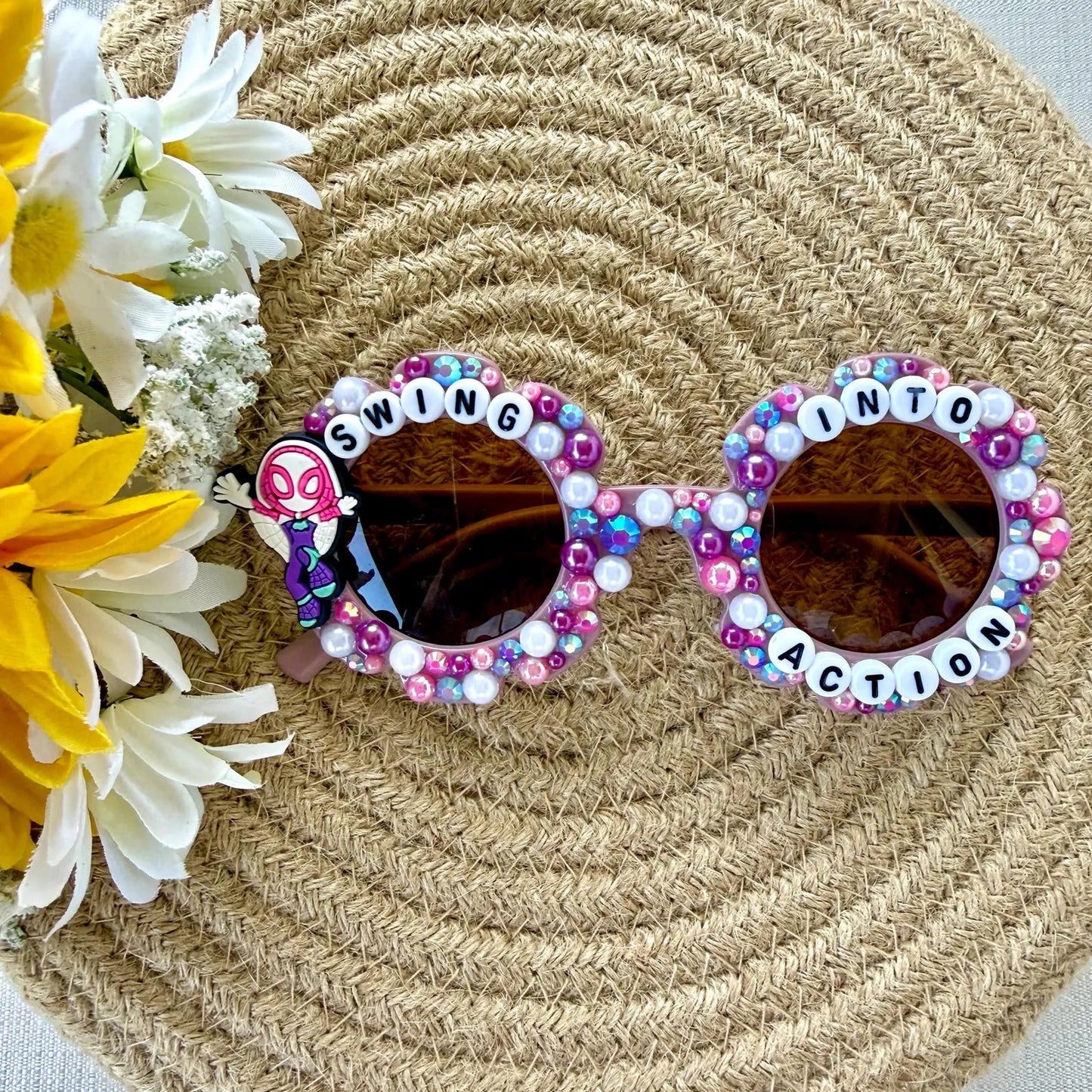Decorated kids purple flower sunglasses with colorful rhinestones and letter beads spelling 'SWING INTO ACTION' on the frames with a Ghost Spider charm on the left side, displayed on a woven straw hat with yellow and white flowers.