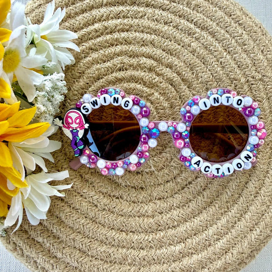 Decorated kids purple flower sunglasses with colorful rhinestones and letter beads spelling 'SWING INTO ACTION' on the frames with a Ghost Spider charm on the left side, displayed on a woven straw hat with yellow and white flowers.