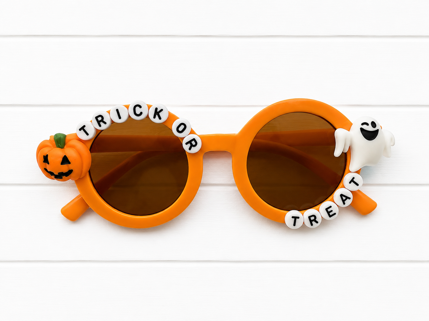 Orange round-frame novelty sunglasses with brown lenses, decorated with a jack-o'-lantern on one arm, a white ghost on the other, and white letter beads spelling 'TRICK OR TREAT' across the top.