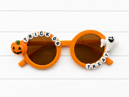 Orange round-frame novelty sunglasses with brown lenses, decorated with a jack-o'-lantern on one arm, a white ghost on the other, and white letter beads spelling 'TRICK OR TREAT' across the top.