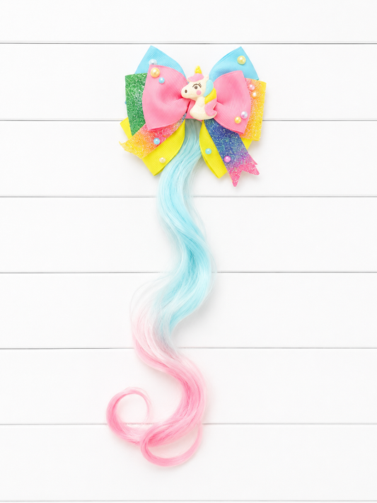 A colorful hair bow accessory with a long wavy pastel tail, featuring a pink and blue glitter bow, a small unicorn charm, and a gradient hair extension that fades from light blue to pink, displayed on a white wood plank background.