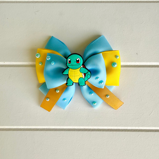 A hair bow featuring light blue and yellow grosgrain ribbon loops with small pearl embellishments, a small Squirtle charm in the center, and orange ribbon tails, displayed on a light gray paneled background.
