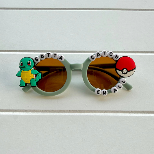 Round sunglasses with blue frames, amber-tinted lenses, a Squirtle charm on the left, and a red-and-white ball charm on the right, with white letter beads spelling 'GOTTA CATCH EM ALL' across the top.