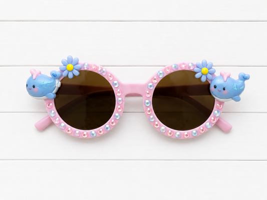 Pink kids round sunglasses with whale and flower charms on either side and blue and pink rhinestones outlining the frames