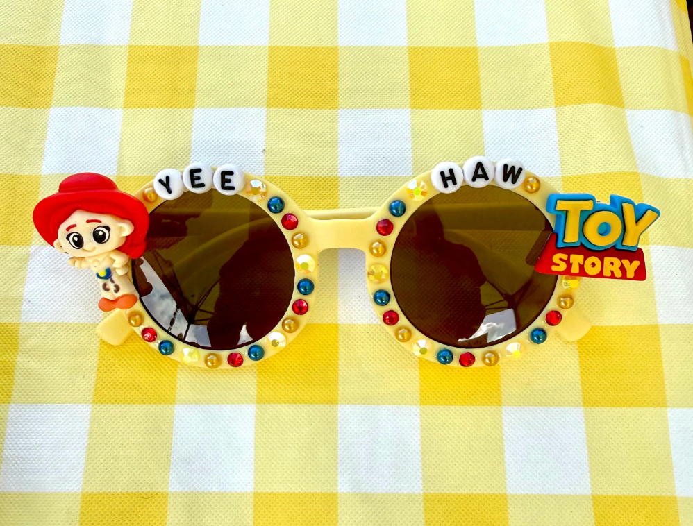 Kids round light yellow sunglasses with Jessie from toy story featured on left, Toy Story logo featured on right, and "Yee Haw" text across the top. There are blue, red, and yellow rhinestones outlining the frames.