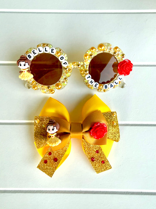 Matching sunglasses and bow. Sunglasses are kids yellow frames with Belle charm on left and red rose charm on right. Gold and yellow pearls and rhinestones and text "BELLE OF THE BALL" outlines the frames. The bow is yellow and gold with Belle and rose charms in the middle.
