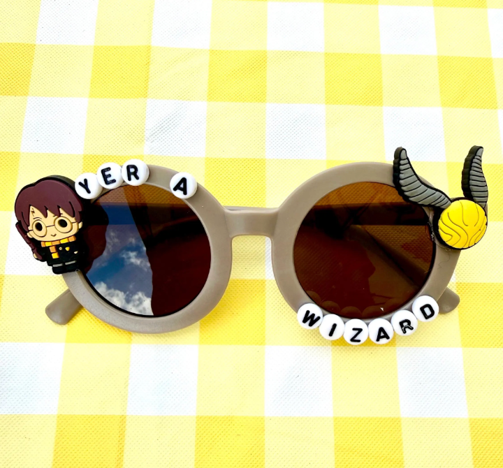 Kids grey round sunglasses with Harry Potter charm on left and Snitch charm on right with "Yer a Wizard" text around the frames.